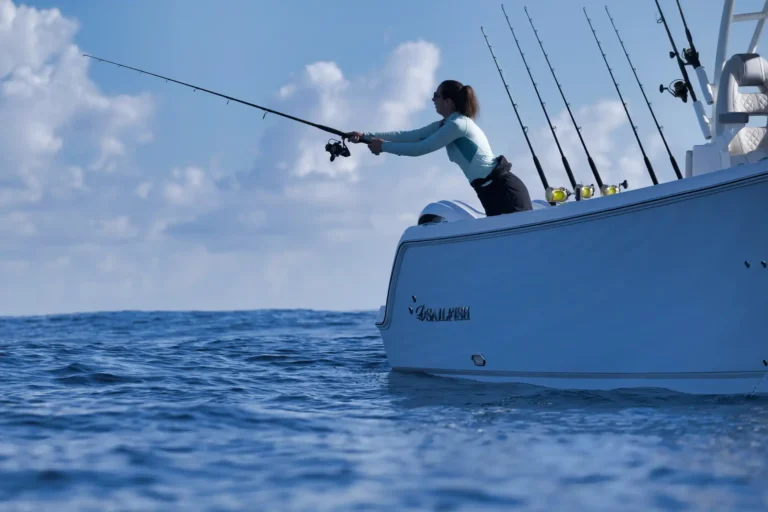 Woman on a white fishing boat casts a rod over the blue ocean, leaning over the side with several fishing poles secured in holders nearby.