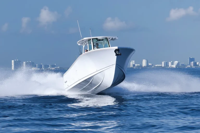 White speedboat skims through blue water with city buildings in the background, creating a spray of waves, sunny sky above.