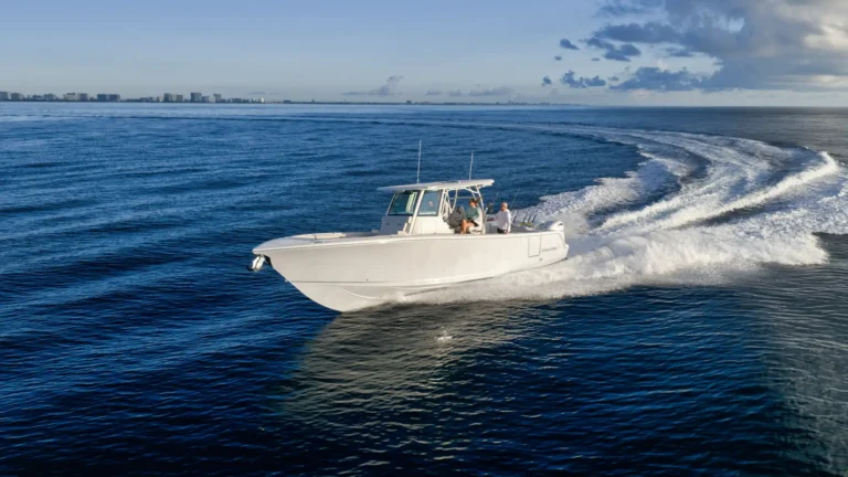 White motorboat cruising across blue ocean with two people aboard, leaving a white wake as a city skyline lines the distant horizon.