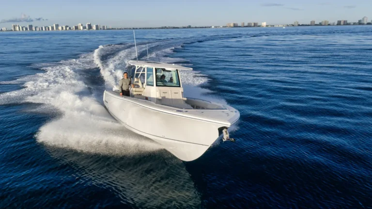White motorboat speeding on blue water, wake behind it, city skyline visible on the horizon with two people aboard.