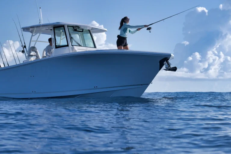 Woman standing on the bow of a white fishing boat casts her line into the blue sea, with a man seated in the cabin behind her.