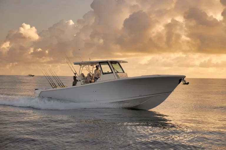 White sportfishing boat speeding across calm ocean at sunset, with multiple fishing rods mounted at the rear and a orange-pink sky reflected on the water.