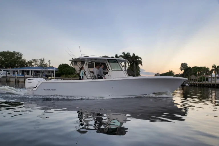White motorboat cruising past a marina with several people on deck, sunset glow in the background and palm trees nearby.