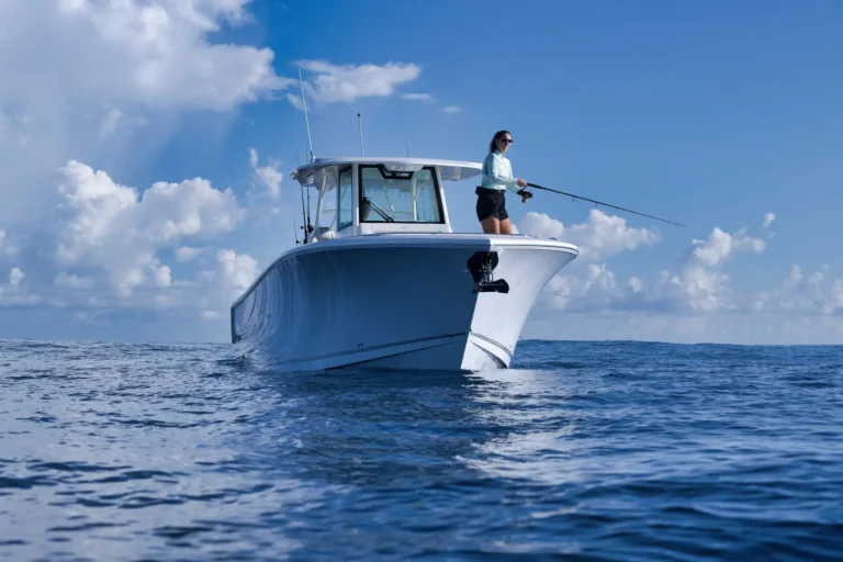 Person standing on the bow of a white boat, fishing rod extended over calm blue ocean under a partly cloudy sky.