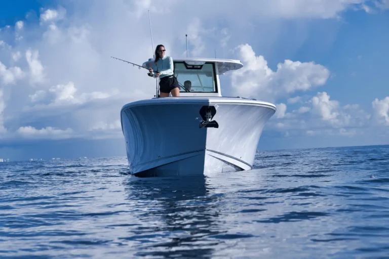 A woman standing on the bow of a white boat, fishing into calm blue water under a cloudy sky