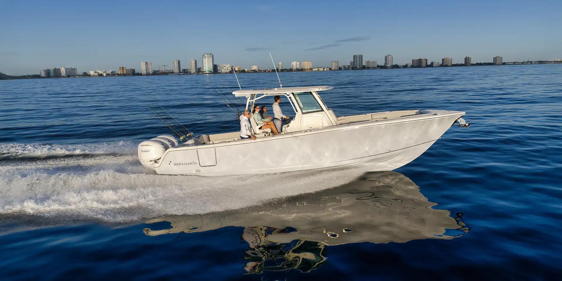 White motorboat with several people cruising on calm blue water near a city skyline at dusk/dawn. Fishing rods on the stern.