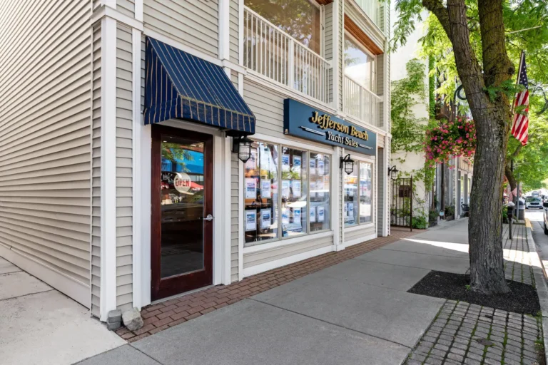 Yacht sales storefront on a sunny street with beige siding, blue striped awning, and large display windows.