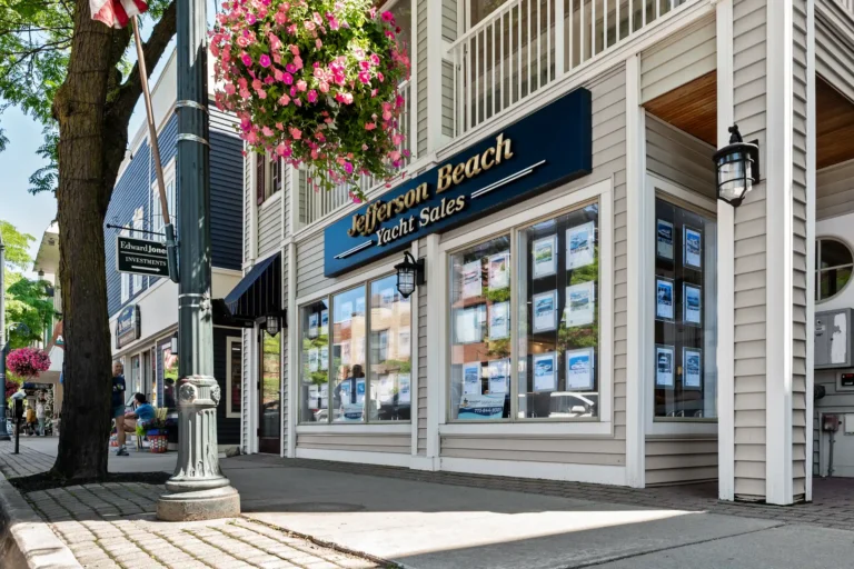 Street view of Jefferson Beach Yacht Sales storefront with a blue sign, large window displays, and a hanging pink flower basket above the entrance.