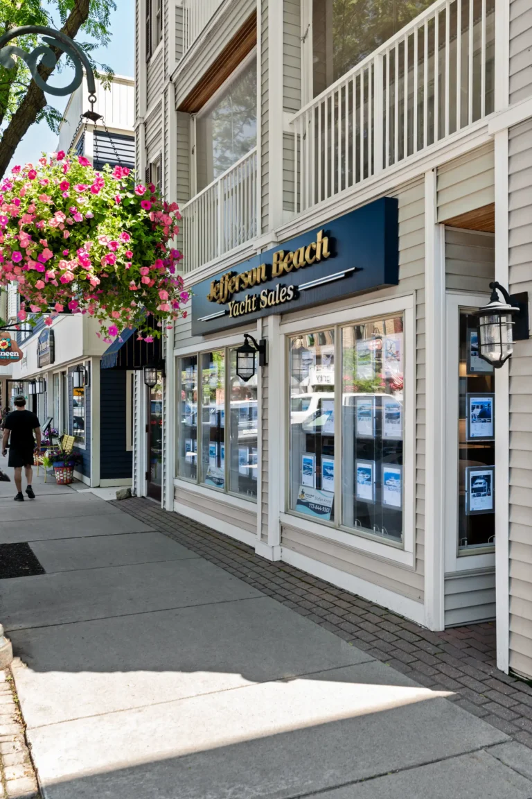 Storefront of Jefferson Beach Yacht Sales with a large blue sign, white siding, and a hanging pink flower basket.