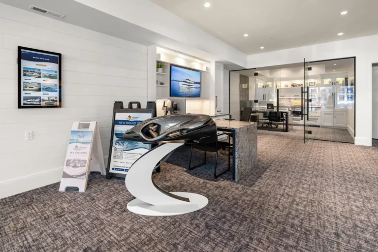 Modern showroom with a sleek boat model on a curved white pedestal, brochure stands, and glass-walled office area in the background.