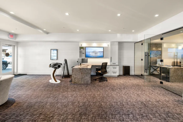Modern office reception area with a marble desk, white walls, and a glass-walled meeting room on the right; monitor shows a boat on screen.] .