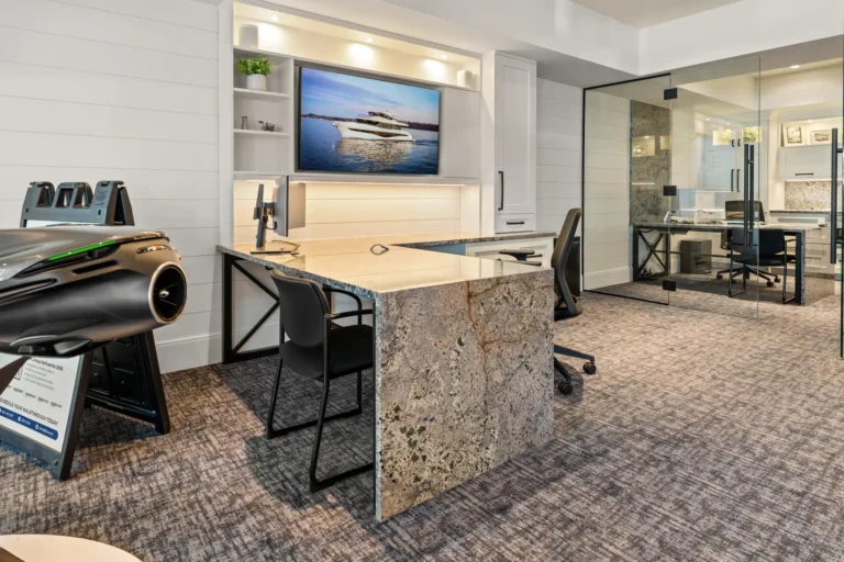 Modern showroom office with a granite island desk, white cabinetry, and a wall-mounted TV showing a luxury yacht on calm water.