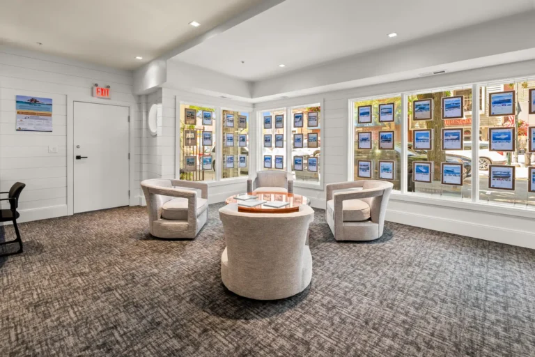 Waiting area of a real estate office: four cream chairs arranged around a glass-topped circular table, with photo displays in the large windows behind.