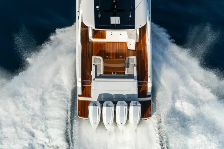 Aerial view of a white luxury yacht from above, cruising through dark blue water with a polished wooden deck and eight outboard engines at the stern leaving a wake behind