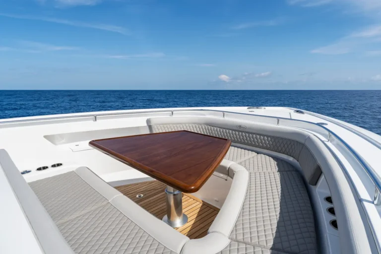 White luxury yacht deck with quilted cream seating around a polished wooden table and a blue ocean horizon above the bow.