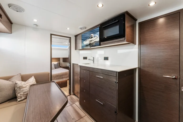 Modern boat galley with dark wood cabinets, white countertop, stainless sink, and built-in microwave above; seating area nearby.