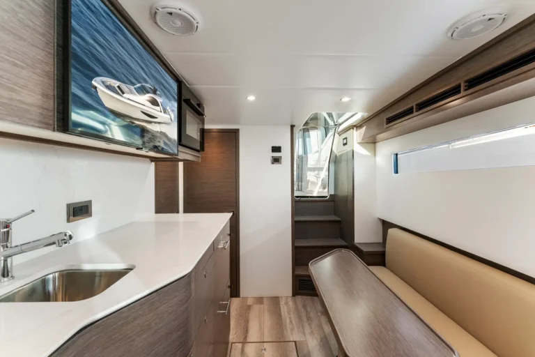 Interior of a modern boat cabin with a kitchenette on the left, a white countertop and sink, and a long beige bench seating along the right side.