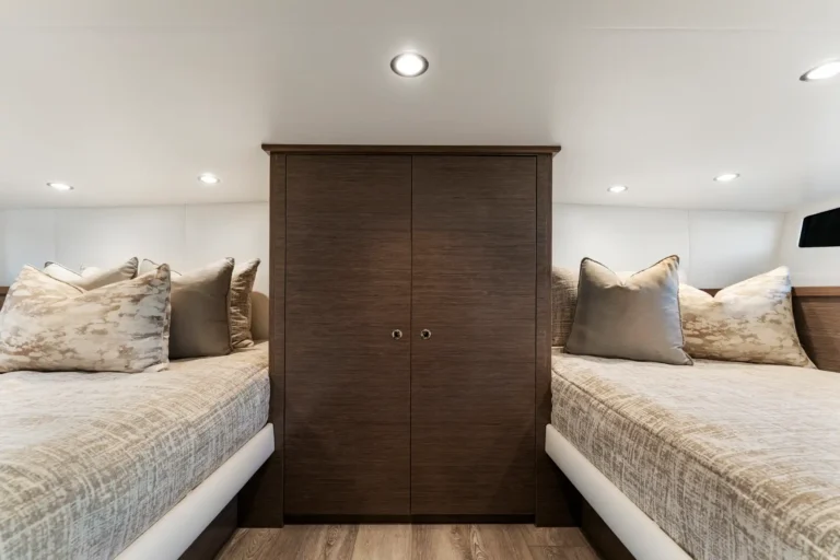 Twin beds with beige patterned bedding flank a central dark wood wardrobe in a cozy cabin bedroom, with recessed overhead lights above.