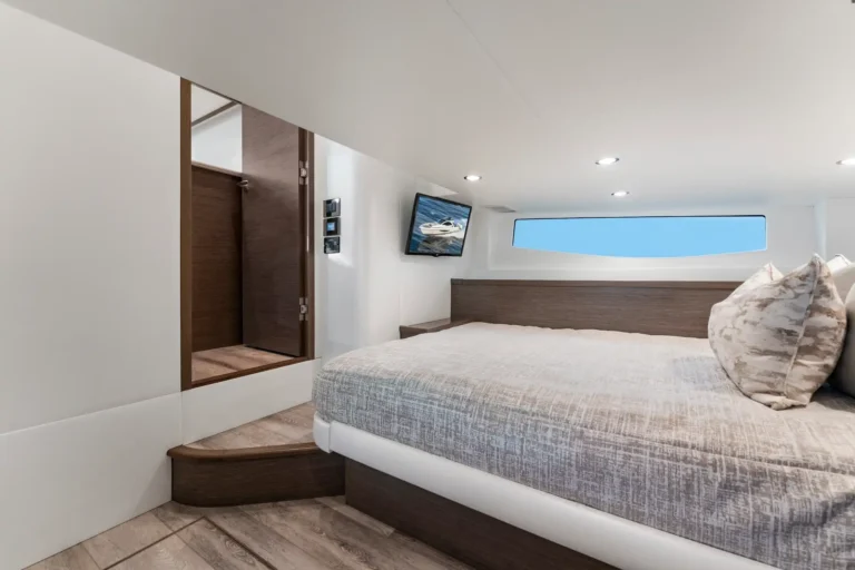Bright yacht cabin with a large bed, wood headboard, gray patterned bedding, and a wall-mounted TV across from the bed.