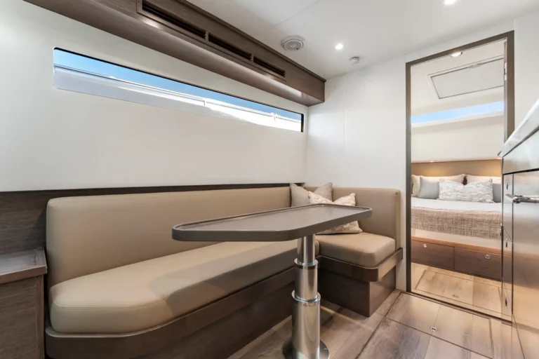Modern boat/yacht interior: beige booth seating with a curved pedestal table and a long horizontal window above. Silvery wood trim and light flooring visible.
