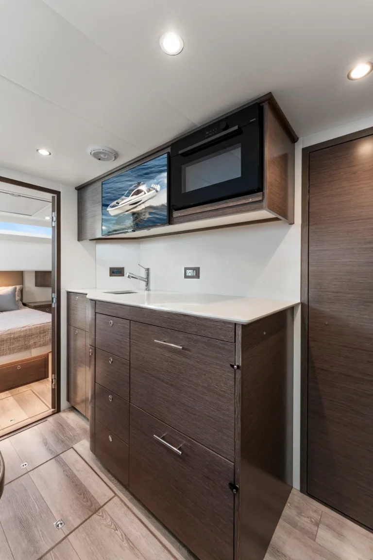 RV kitchen with dark wood cabinetry, white countertop, sink, and built-in microwave; wall TV shows a speedboat image.