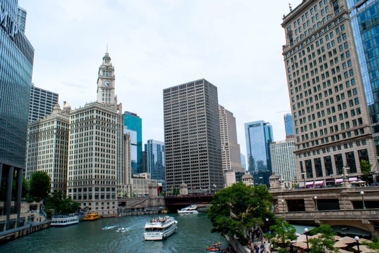 Wide view of a city riverfront with tall office buildings lining both banks and boats cruising on the water.