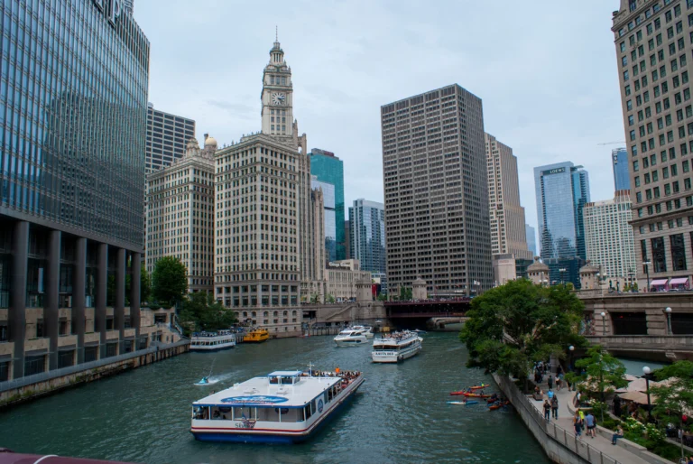 Downtown Chicago riverfront with tour boats and tall office buildings lining the riverwalk under a pale blue sky client-side shadows on glass façades