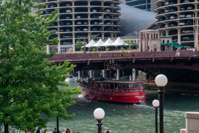 Red sightseeing boat on the Chicago River under a bridge, with a State Street sign and a multi-level parking garage in the background.
