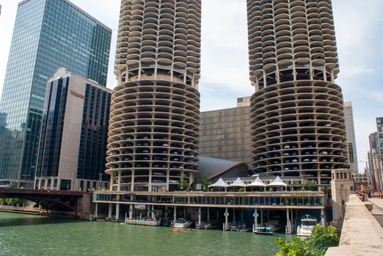 Twin circular-balcony Marina City towers along the Chicago River, with a glass office building nearby and a riverfront promenade in the foreground.