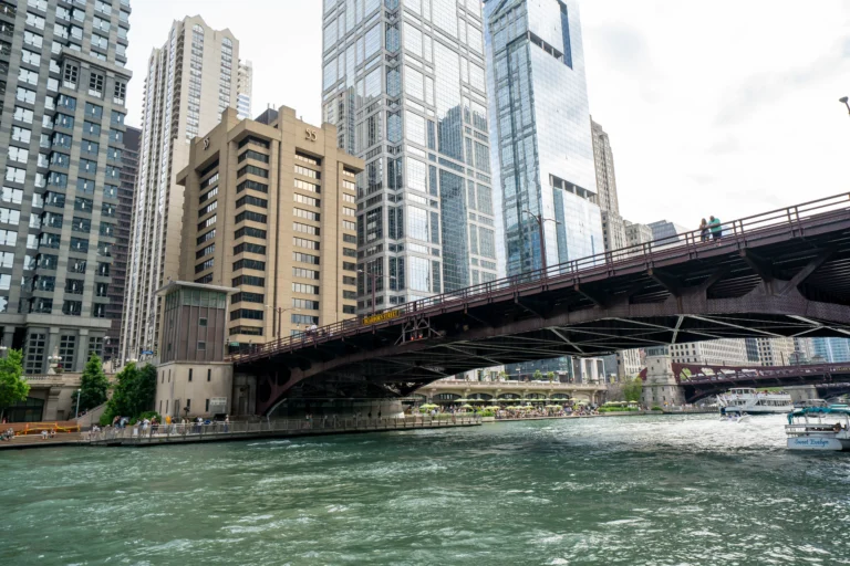 City skyline with tall glass buildings along a river and a brown steel bridge with pedestrians crossing on the right.