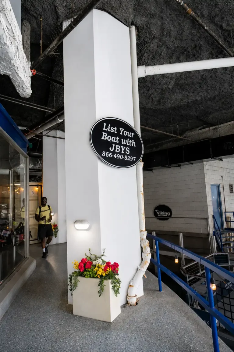 Indoor showroom entrance with a circular JBYS sign on a white pillar and a flower-filled planter at its base.