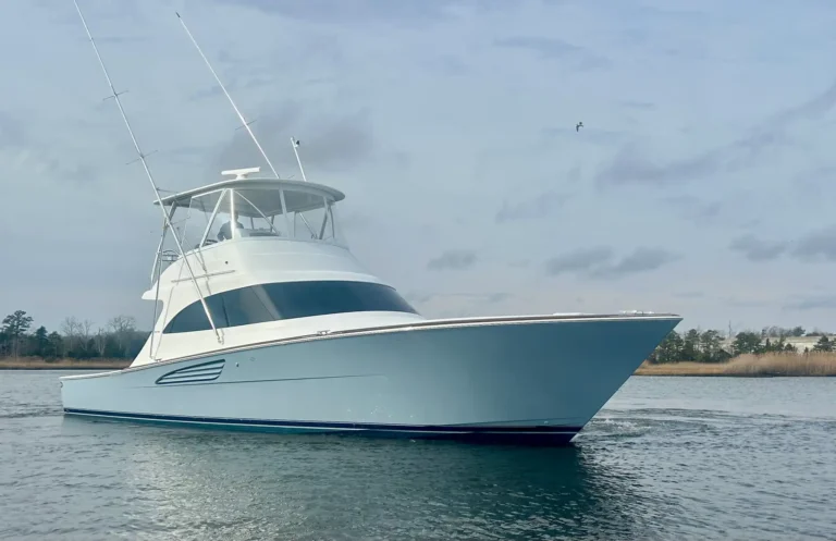 White yacht with a blue hull cruising on calm water, tall antennas rising from the deck, shoreline in the distance.