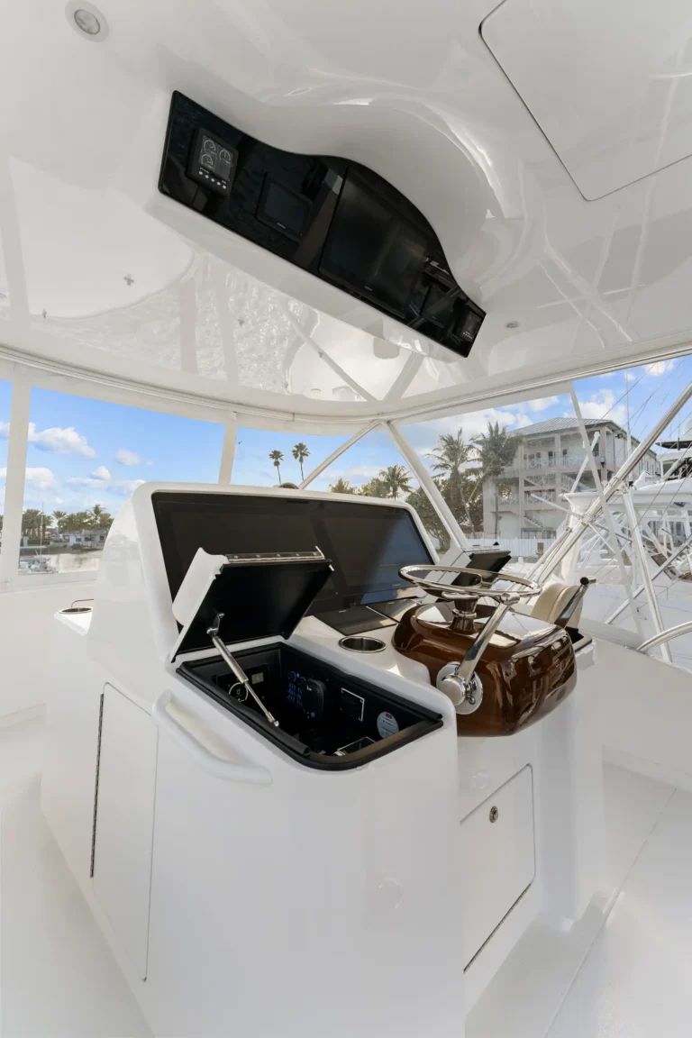 Yacht helm console with open electronics hatch, showing navigation screens and controls beside a wooden steering wheel on a glossy white deck.