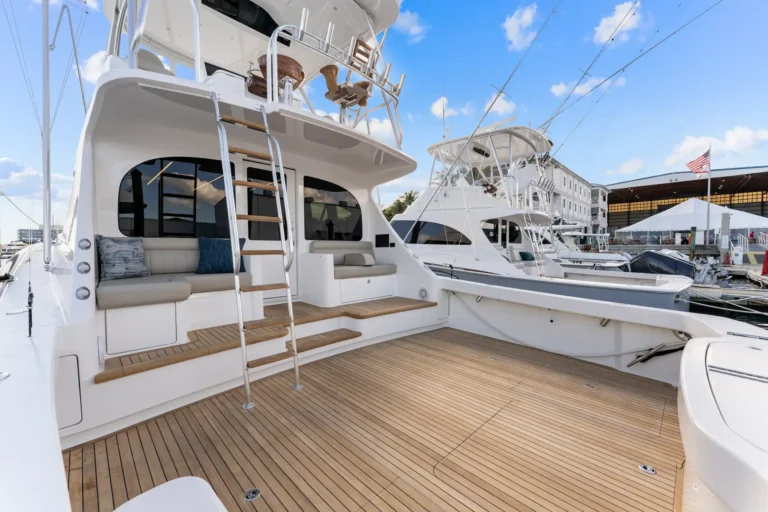 Teak-decked aft deck of a white luxury yacht with seating and a ladder at a marina.