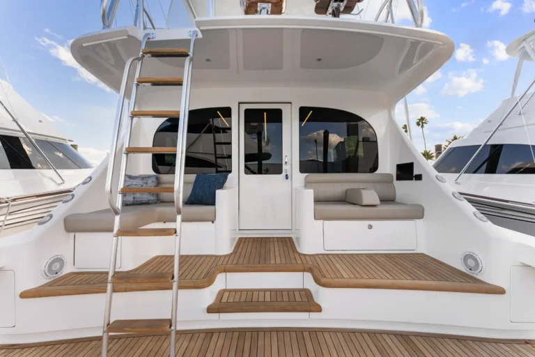 Rear view of a luxury yacht’s stern with a metal ladder, teak steps, and cushioned seating on a teak deck under a blue sky.