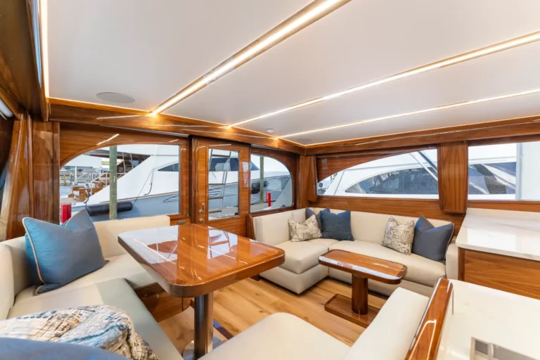 Luxurious yacht salon with cream sofas, blue cushions, polished wood tables, and warm ceiling lighting.