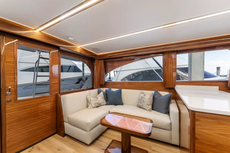 Luxurious wood-paneled yacht salon with a cream sectional sofa and pink marble coffee table, large windows showing another yacht outside.