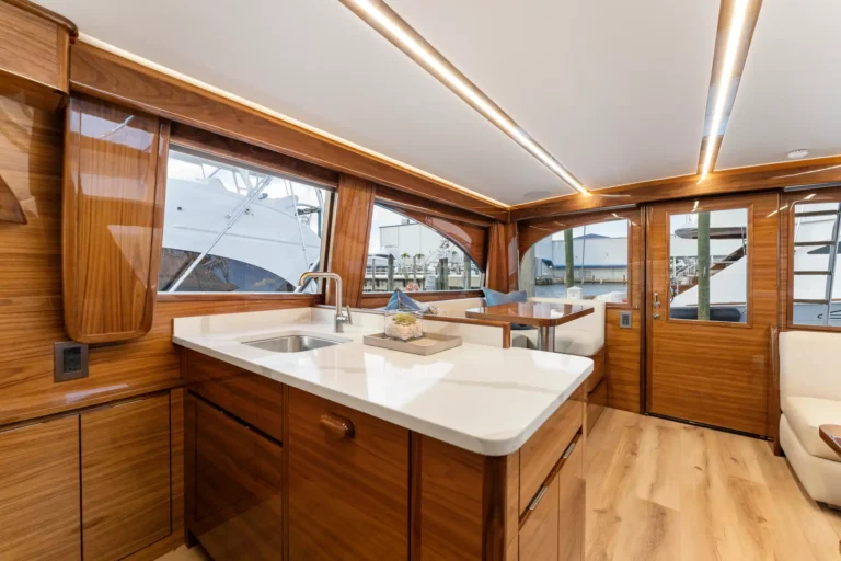 Luxurious teak wood yacht galley with a white countertop, sink, and large windows showing the marina outside.