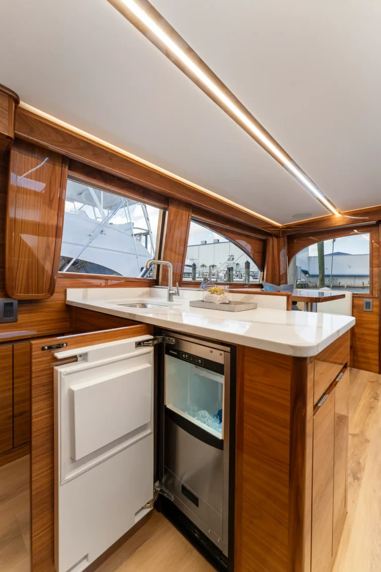 Wood-paneled yacht galley with white countertop, open fridge, and built-in dishwasher beneath a bright ceiling light; windows show marina outside