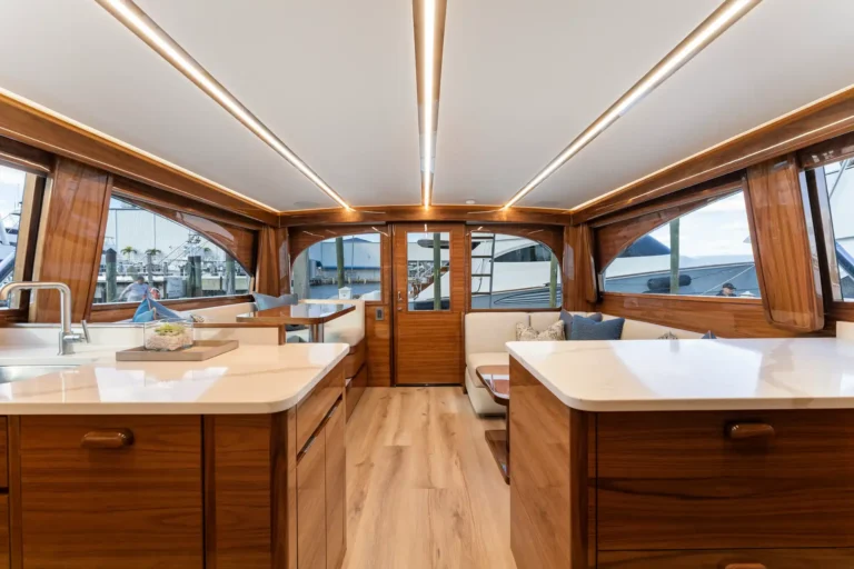Luxurious wooden yacht cabin with white countertops, cream seating, and large windows overlooking a marina.