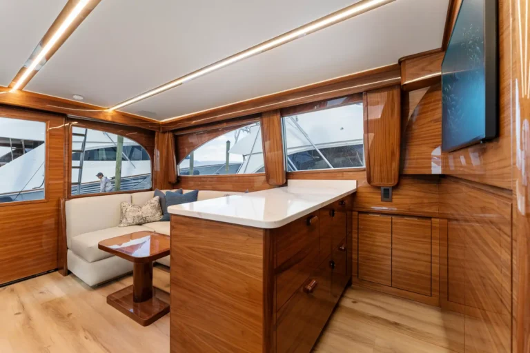 Teak wood yacht cabin with cream seating, white counter, and built-in cabinets along glossy wood walls, illuminated by strip lighting.