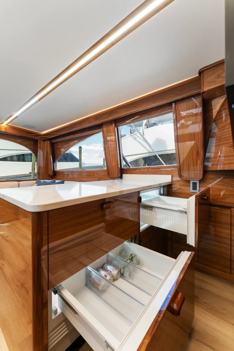 Wooden yacht galley with white countertop, open fridge drawers showing drinks, and warm wood cabinetry along a windowed wall.