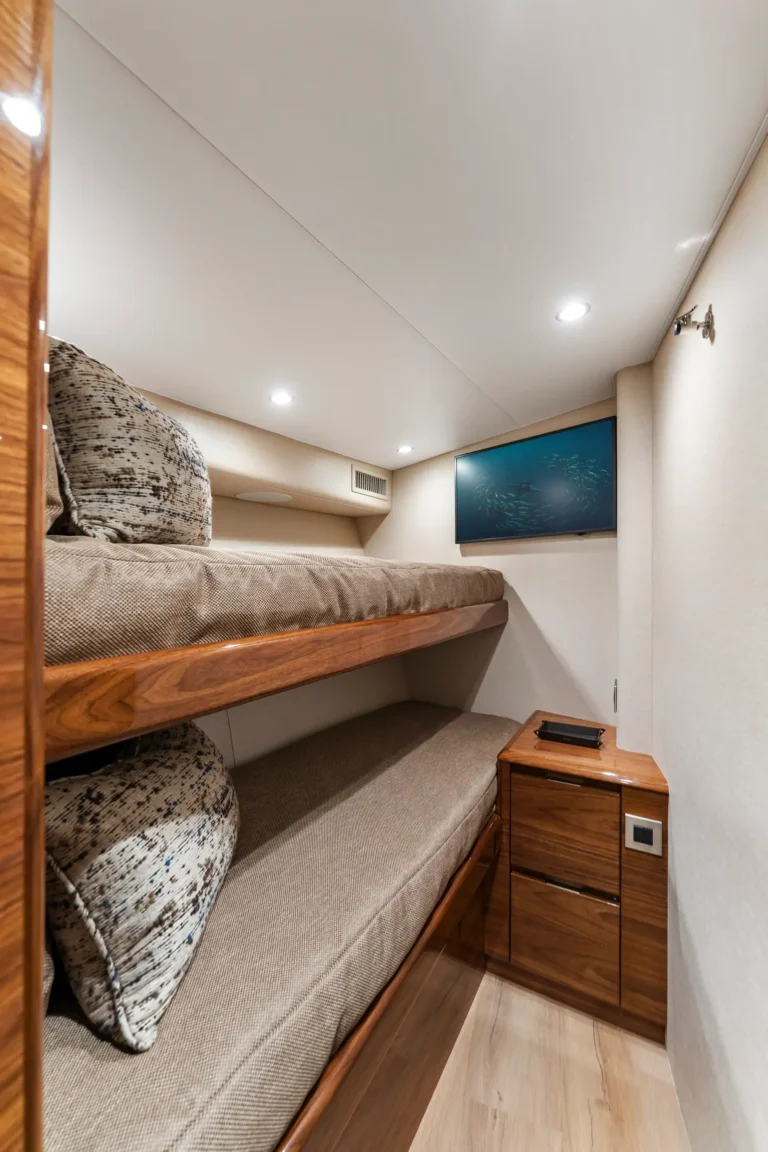 Compact bunk cabin interior with a top bunk, beige upholstery, and a wood nightstand; a TV mounted on the wall opposite the bed.