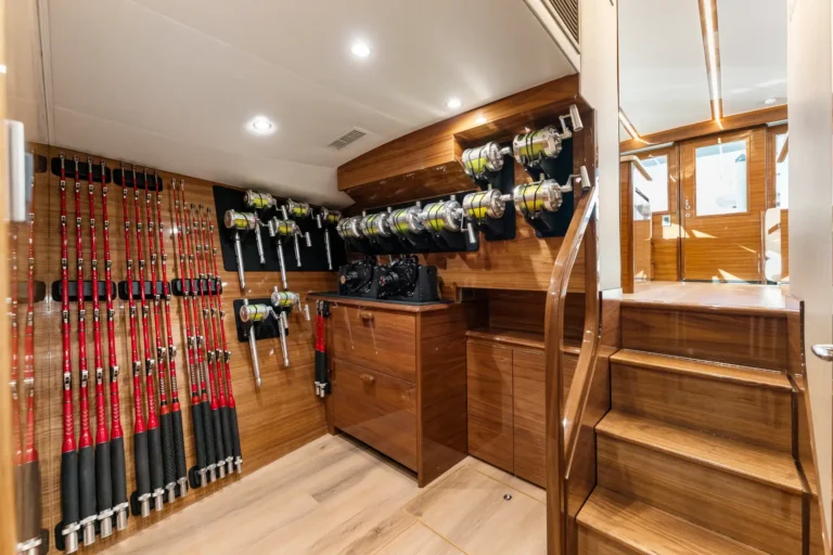 Organized fishing rod storage area aboard a wooden interior yacht, with rods lined on left and reels mounted on wall to the right of a staircase.