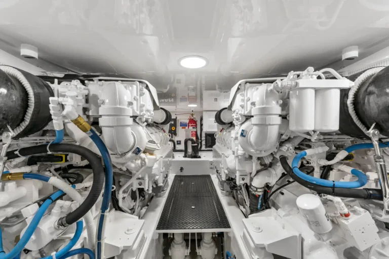 Twin white marine diesel engines in a compact engine room, with blue hoses and hoses, central walkway, and control panels in the back.