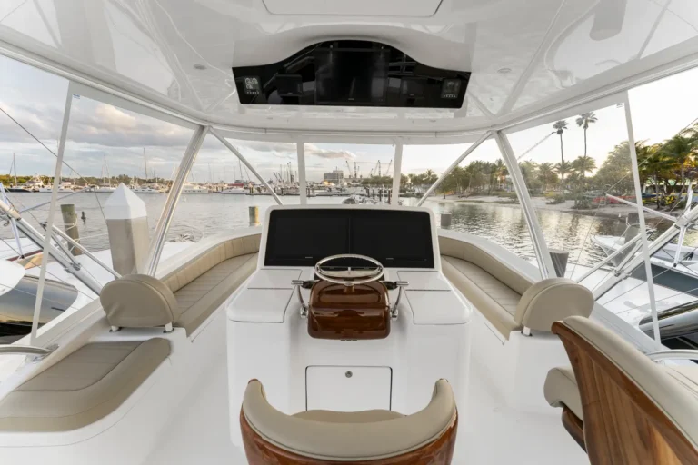 Cockpit of a luxury yacht with beige leather seating, dual screens, and a wooden helm, marina visible outside the windows.