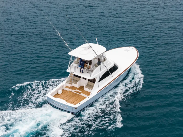 Top-down view of a white motor yacht cruising through deep blue sea with passengers on the deck and a foamy wake behind it.