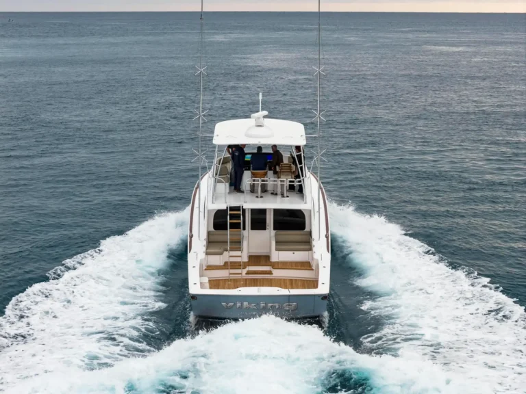 Rear view of a white motor yacht cruising the open sea, wake foaming at the stern and passengers visible on deck behind the cockpit (Viking branding on hull).”,