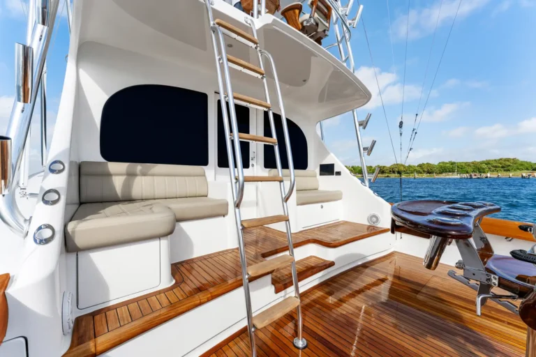 Rear cockpit of a luxury yacht with teak deck, beige seating, and a stainless steel ladder to the upper deck on a sunny day by calm water.