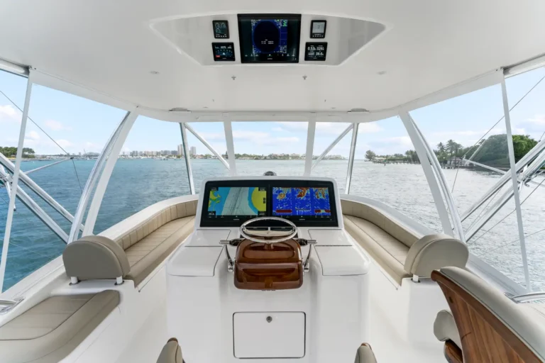 Cockpit of a white yacht with navigation screens and steering wheel, blue water outside and a distant shoreline visible through the windows